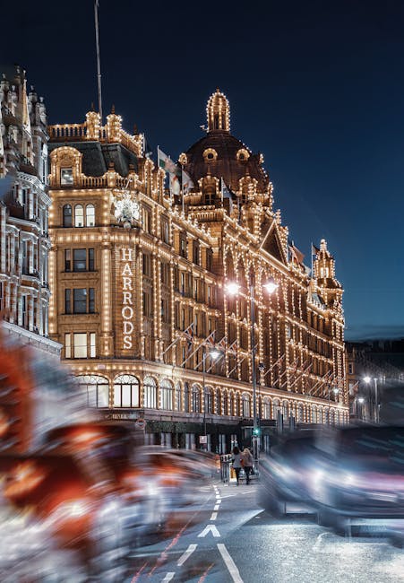 Nighttime exterior view of a historic building illuminated with festive string lights, showcasing ornate architectural details and large windows, with a vertical sign reading 'HARRADS' along the facade. The street in front is busy with moving vehicles, resulting in motion blur effects, and a few pedestrians walking on the sidewalk. The scene captures the lively atmosphere of Knightsbridge, in the vicinity of high-end shopping and homes, with the glowing lights reflecting the vibrancy of the area. The environment suggests urban transportation and the movement of belongings, relevant to house removals and furniture transport services provided by [COMPANY_NAME], especially during home relocation or packing and moving activities. This visual emphasizes the importance of professional moving logistics within a bustling cityscape, supporting the topic of avoiding hidden removals charges in Knightsbridge moves.