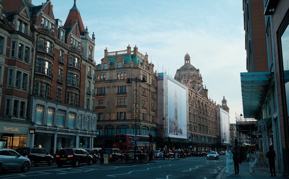 A busy urban street scene in Knightsbridge, featuring historic and modern buildings with detailed facades, large windows, and decorative architectural elements. Several black and white cars are parked along the street, with some vehicles in motion. Pedestrians are walking on the sidewalk, and a red double-decker bus is visible in the background. On the right side, there is a building under renovation with scaffolding and a large advertising banner hung between ornate street lamps. The scene is captured during daytime with clear weather, and natural light illuminates the street, highlighting the intricate design of the surrounding architecture. The environment suggests a location suitable for professional home relocation and furniture transport, with the focus on the urban setting and street-level activity that may be associated with moving logistics, loading, and unloading activities coordinated by house removals specialists like [COMPANY_NAME].
