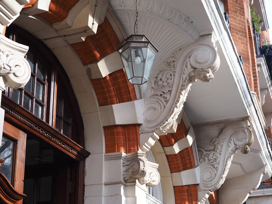 Close-up view of an ornate residential building facade with decorative white plasterwork featuring intricate scrollwork and floral motifs supported by detailed corbels. The building has a large arched window framed with dark wood, partially open, revealing a glimpse of interior structures. Above the window, a traditional outdoor lantern with a glass enclosure hangs from a chain, illuminating the entryway area. Surrounding the architectural details are red brick accents that contrast with the white decorative elements. The scene is set outdoors in daylight, with natural light highlighting the craftsmanship of the ornate trim and the texture of the brickwork, reflecting the type of property often involved in home relocation or furniture transport services. This detailed architectural image is relevant for discussing common access problems for Knightsbridge removals and their solutions, as managed by companies like removalcompaniesknightsbridge.co.uk.