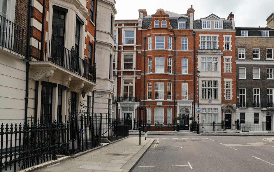 The image depicts a street scene in Knightsbridge featuring a row of multi-storey residential buildings with classic London architectural styles, including ornate window frames and brick facades. In the foreground, there are black wrought iron fences bordering the properties, with some small potted plants visible near the entrance gates. The street is relatively quiet with minimal vehicle or pedestrian activity, and a few yellow parking restriction lines are painted on the asphalt. The pavement is made of light-colored stone slabs, and the surroundings suggest a well-maintained, affluent neighbourhood. This scene provides context for home relocation or furniture transport, illustrating the typical environment where house removals by companies like [COMPANY_NAME] are conducted, especially when navigating tight urban access points for efficient packing and loading processes during house moves in Knightsbridge.