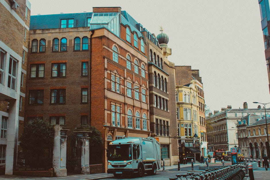A blue and white removal vehicle is parked on a city street in front of a row of multi-storey buildings with a variety of architectural styles, including brick and ornate facades. The vehicle is positioned near a curb and adjacent to a loading area where removal and packing activities are likely taking place. Nearby, several large cardboard boxes, some wrapped in plastic and others open to reveal packing materials, are arranged on the pavement, with a few being carried or loaded onto the van. Trolleys, straps, and moving blankets are visible, indicating a professional home relocation process managed by [COMPANY_NAME]. The scene captures a typical urban moving operation involving furniture, packed boxes, and transportation equipment, consistent with the requirements for house removals, transportation of furniture, and packing services. The environment includes surrounding buildings, street fixtures, and a bright, overcast sky, typical of a busy London neighbourhood such as Knightsbridge, where careful planning for removals adhering to local council rules is essential.