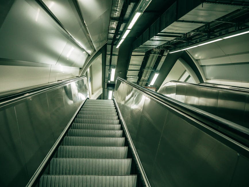 A downward view of an escalator inside a modern indoor setting, with metal side panels and a ribbed rubber surface on the steps. The escalator is positioned between curved, smooth, metallic walls illuminated by overhead lighting, and is surrounded by structural elements including visible pipes and steel beams, indicating an underground or station environment. The bright lighting highlights the clean, polished surfaces, and the escalator appears to be in the process of transporting passengers or items between floors. This setting reflects the kind of environment where a professional removals company like [COMPANY_NAME] might coordinate the moving of furniture, boxes, or appliances in an efficient, well-structured manner during home relocation or transit within multi-level buildings, such as stations or commercial complexes, with a focus on smooth loading and unloading processes.