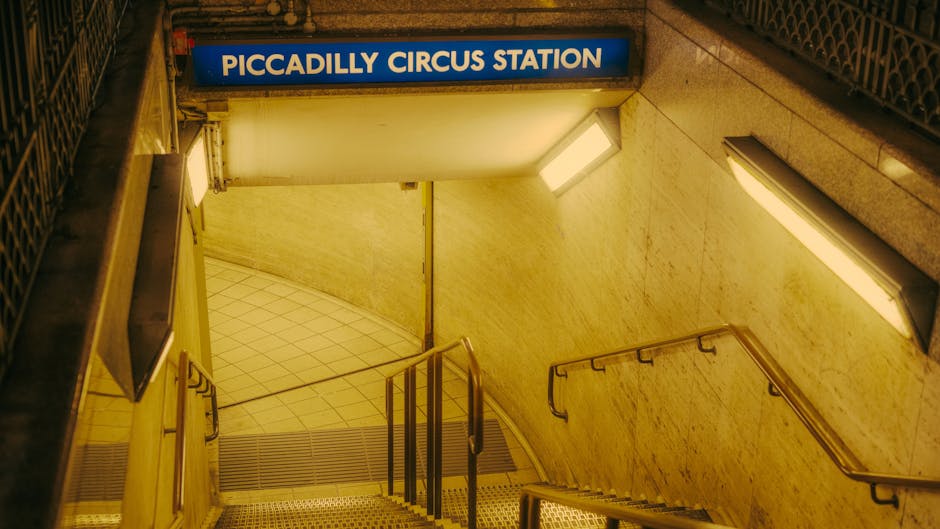 The image shows a downward view of the entrance to Piccadilly Circus Station in London, with a blue and white sign displaying the station name at the top. The staircase is constructed from metal and concrete, with black handrails on either side for support. The stairs are metallic and textured, leading down into the station, and there are two large rectangular fluorescent lights on the beige, textured tiled walls on either side, providing illumination for the descent. The area appears to be part of a housing or commercial building's underground station access, designed for pedestrian movement. This setting is relevant for house removals and moving services, such as those provided by [COMPANY_NAME], where careful handling of furniture and boxes during the loading process is essential when navigating tight access points like underground station staircases, ensuring safe and efficient transportation of belongings during a home relocation or furniture transport.