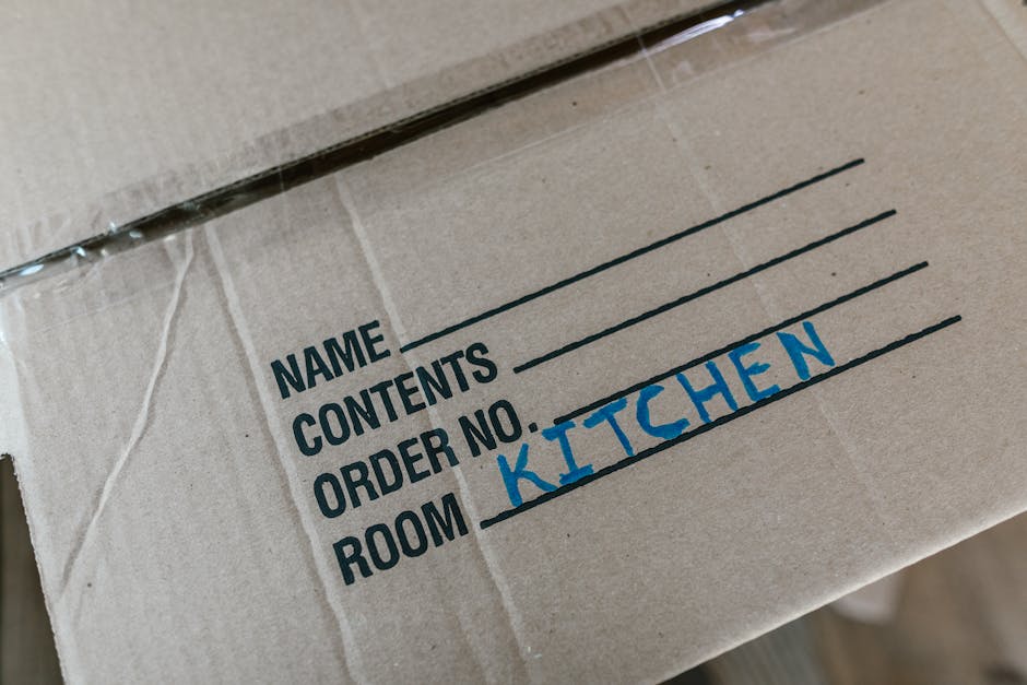 Close-up image of a cardboard moving box labeled with a printed and handwritten section, showing the printed words 'NAME', 'CONTENTS', 'ORDER NO.', and 'ROOM'. The 'ROOM' section has been filled in with the handwritten word 'KITCHEN' in blue ink. The box is sealed with clear packing tape along the top edge and is situated inside a property, likely near an entrance or storage area. The environment appears well-lit, emphasizing the details on the box. This image exemplifies proper packing identification during a home relocation, highlighting the importance of clear room labeling to facilitate efficient furniture transport and packing and moving processes, which are relevant to professional removals services such as those offered by [COMPANY_NAME].