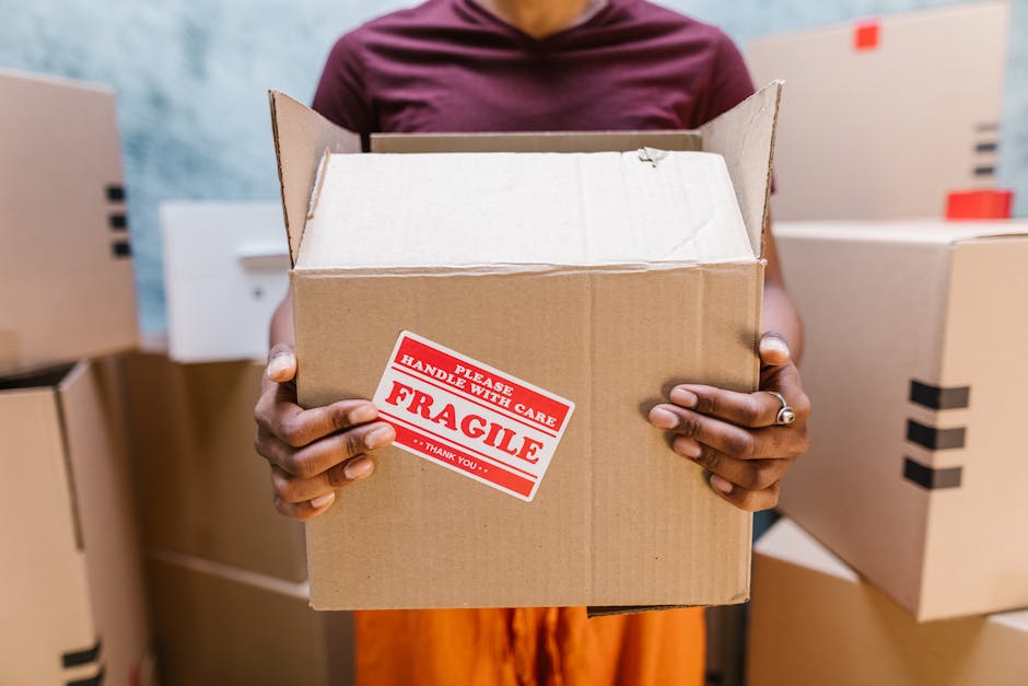 A person holding an open cardboard box with a red and white 'Fragile Handle with Care' sticker visible on the front. The person’s hands, wearing a silver ring, grip the box securely, while their torso is partially visible, clothed in a maroon top. Surrounding the individual are several other cardboard boxes, some sealed with black tape, stacked in what appears to be a storage or packing area, with a light-colored textured wall in the background. The scene is well-lit with natural or artificial light, emphasizing the focus on safe packing practices during home relocation. The image illustrates careful handling of fragile items, relevant to professional removals services such as those offered by [COMPANY_NAME], emphasizing correct packaging to prevent damage during furniture transport and loading processes.