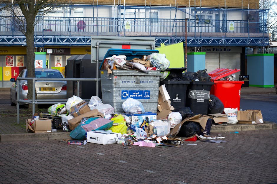 An outdoor urban scene showing overflowing waste and recycling bins and surrounding litter, including cardboard boxes, paper, plastic bags, and packaging materials on the pavement near parked cars. The waste containers are of various sizes and colors, with one grey mixed paper and cardboard bin, a black bin for general waste, and a red bin, all positioned close to a metal fence and a row of parked vehicles. Behind the bins, there is a building with storefronts and signage, indicating a commercial area. The scene is lit by natural daylight. This image relates to waste disposal regulations relevant to house removals and moving services provided by [COMPANY_NAME], emphasizing the importance of proper waste management during home relocation or disposal processes, aligned with the context of [PAGE_TITLE].