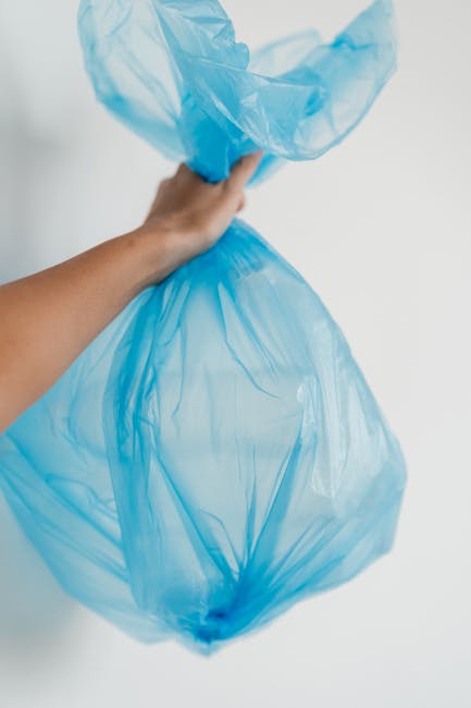 A person's hand holding a large, semi-transparent blue plastic waste bag, with folded edges and slight creases, suspended in front of a plain white background. The bag appears to be filled with waste material, suitable for disposal during house removals or home relocation processes. The hand grips the bag at the top, indicating an active waste disposal or packing task. The image highlights the importance of responsible waste management in moving services provided by companies such as removalcompaniesknightsbridge.co.uk. This scene reflects the packing and waste disposal stage involved in moving logistics, emphasizing proper separation and handling of waste, cardboard boxes, and packing materials typical in house removal operations.