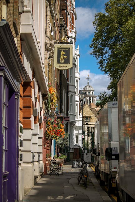 A narrow street scene in Knightsbridge, London, featuring historic buildings with ornate facades, balconies decorated with vibrant flower boxes, and an overhead sign for a pub named 'Youngs' hanging from a wrought iron bracket. A partially visible moving truck with a reflective, semi-transparent side panel is parked on the right, alongside a bicycle leaning against a railing. In the background, a domed historic cathedral with classical columns and a clock tower can be seen under a partly cloudy sky, indicating a busy area prepared for a house relocation or furniture transport. The lighting suggests daytime with natural sunlight illuminating the scene, capturing elements typical of an urban move managed by a professional removals company like [COMPANY_NAME].