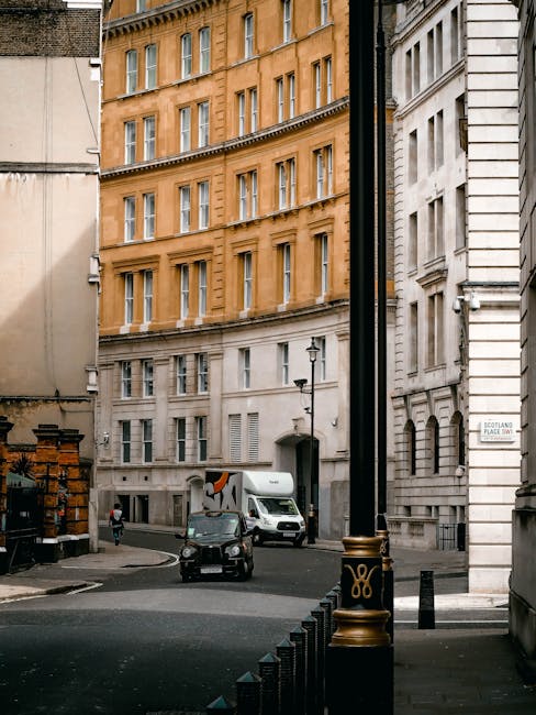 Photograph of a narrow street in Knightsbridge with multi-storey buildings featuring large windows and classical architectural details, including cream and beige facades. In the foreground, a black, gold-accented street bollard is visible, along with a black pole and a portion of a white building on the right. A small black vehicle, likely a compact car, is parked near the curb, while a white moving van with a partially visible logo is positioned further into the street, indicating a home relocation or furniture transport process. Behind the van, a person in dark clothing is walking near the motorized vehicles. The scene is outdoors during daylight with overcast weather, suitable for professional removals services by companies like [COMPANY_NAME], engaged in packing, loading, or transporting household furniture and belongings within this urban environment.