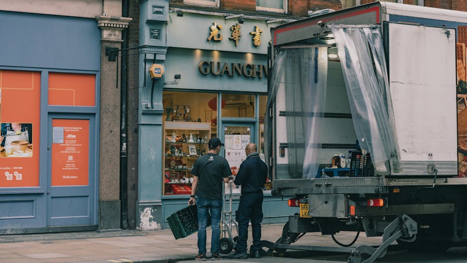 A narrow street scene in Knightsbridge, London, featuring historic buildings with ornate facades, balconies decorated with vibrant flower boxes, and an overhead sign for a pub named 'Youngs' hanging from a wrought iron bracket. A partially visible moving truck with a reflective, semi-transparent side panel is parked on the right, alongside a bicycle leaning against a railing. In the background, a domed historic cathedral with classical columns and a clock tower can be seen under a partly cloudy sky, indicating a busy area prepared for a house relocation or furniture transport. The lighting suggests daytime with natural sunlight illuminating the scene, capturing elements typical of an urban move managed by a professional removals company like [COMPANY_NAME].