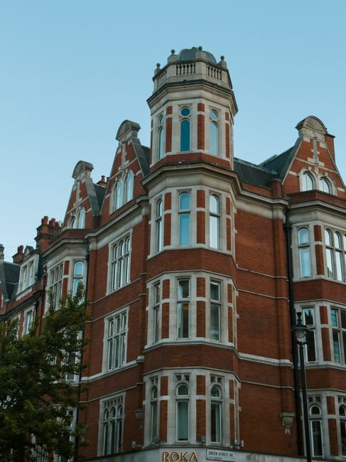 A multi-storey red brick building with white window frames and decorative stone detailing, typical of Victorian architecture, located on a street with a clear blue sky background. The building features a prominent turret with a rounded, crenellated roof and ornate railings, along with several large sash windows and gabled sections. Part of the street scene includes a black lamppost beside the building. This image provides a visual context for properties involved in house relocations or moving services, such as those offered by removalcompaniesknightsbridge.co.uk, which specialise in furniture transport, packing, and loading processes for home relocation projects.