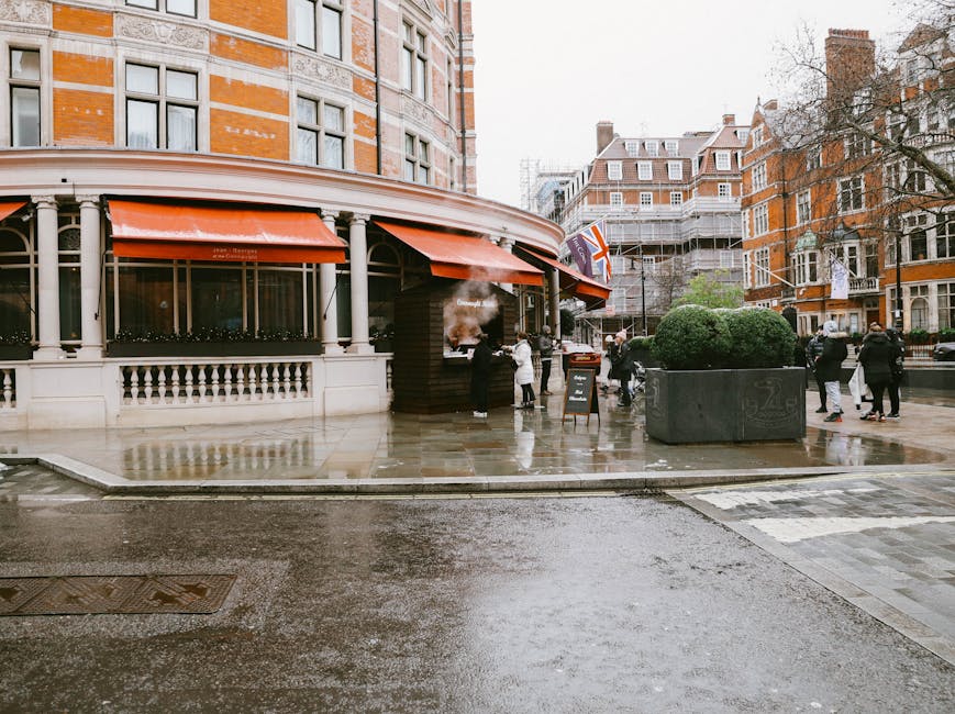 A street scene in a city featuring a corner building with a restaurant or café, having red awnings and decorative railing, on a wet, rainy day. There are several pedestrians walking on the sidewalk, some carrying bags or parcels. A small outdoor seating area is visible, with a chalkboard sign placed beside it. In the background, multi-story brick and stone buildings with various windows and architectural details can be seen. The street is moist, reflecting light from the overcast sky, and a large planter with green shrubbery is positioned near the building. No moving equipment or furniture is visible in the image, but the setting is typical of urban environments where professional removal services, such as those provided by [COMPANY_NAME], often assist with home relocation and furniture transport during house removals in central London.