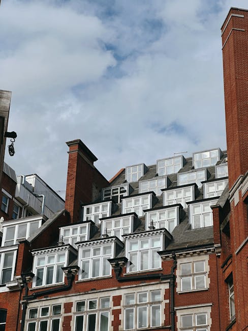 Photograph of the upper floors and roof of a red brick residential building with multiple white-framed bay windows and dormer windows. The building features decorative brickwork and chimney stacks, with a partial view of a lantern-style exterior light fixture on the left side. The overhead sky is partly cloudy with patches of blue, giving natural lighting to the scene. This setting reflects the typical architecture found in high-end London residence buildings, pertinent to house removals and home relocation services. The image emphasizes the external structure and window arrangements, which are relevant when planning furniture transport and packing considerations for a SW1X penthouse move, as managed by companies like [COMPANY_NAME], experienced in detailed removal logistics.