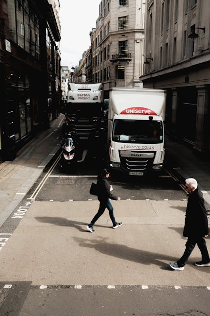 A street scene in Knightsbridge featuring two white removal trucks with branding from Uniserve and Becton Transport parked on a narrow road. The trucks are positioned closely to each other, partially blocking the street, with a motorcycle parked adjacent to them. In the foreground, two pedestrians are crossing the street; a woman in casual clothing carrying a black bag and an older man with white hair dressed in dark clothing. The surrounding buildings are multi-story, with large windows and traditional stone facades, typical of historic London architecture. The scene is lit by natural daylight, and the environment suggests a busy urban setting suitable for home relocation or furniture transport services by companies like [COMPANY_NAME], supporting last-minute moves or packing and loading operations involved in house removals.