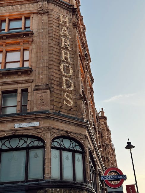 A close-up view of a historic multi-storey building with ornate architectural detailing, featuring a large vertical sign spelling 'FURNITURE' in illuminated letters attached to the corner façade. The building has large, curved glass windows on the ground floor, with decorative ironwork and stonework details. The sky is clear with a few clouds, and a street lamp and an underground station sign are visible in the foreground. This scene depicts the exterior of a location where furniture transport and home relocation services, such as those offered by [COMPANY_NAME], may be involved in house removals and moving logistics in Knightsbridge.
